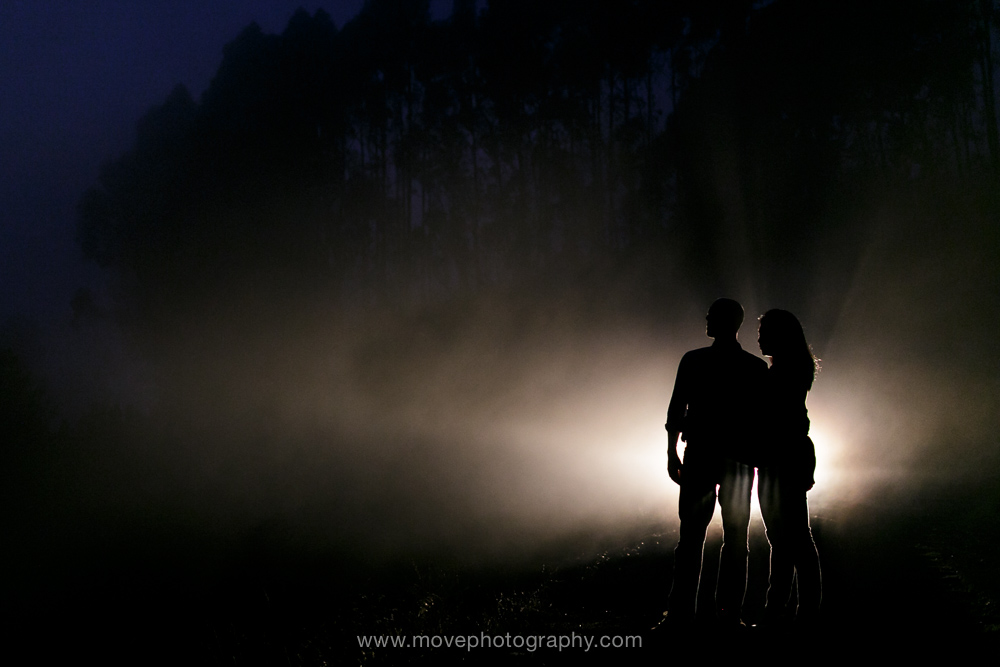 A fiancée and her fiance stand in the glow of their car's headlights in the Oakland hills at twilight, in front of eucalyptus and the blue glow of the sky, after their Rockridge engagement session.
